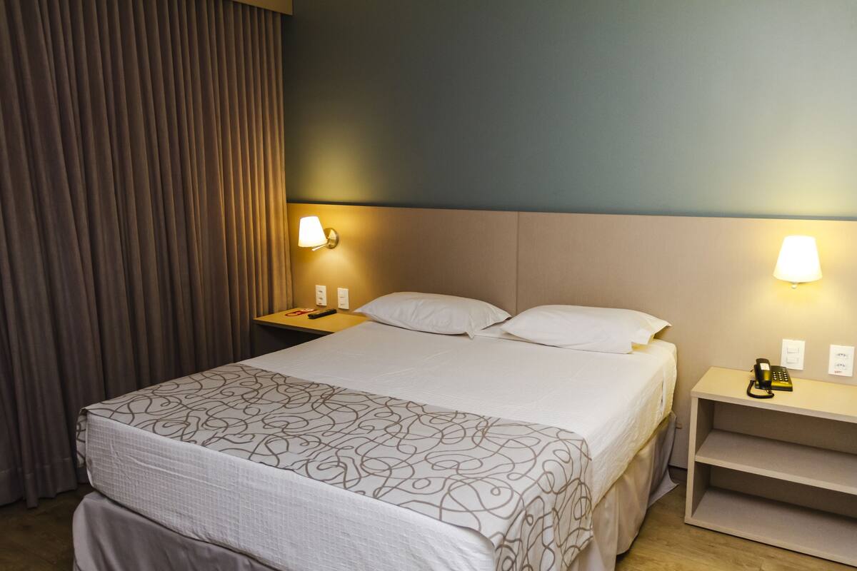 A hotel room with a bed, two bedside tables, a lamp, a phone, and a curtain.