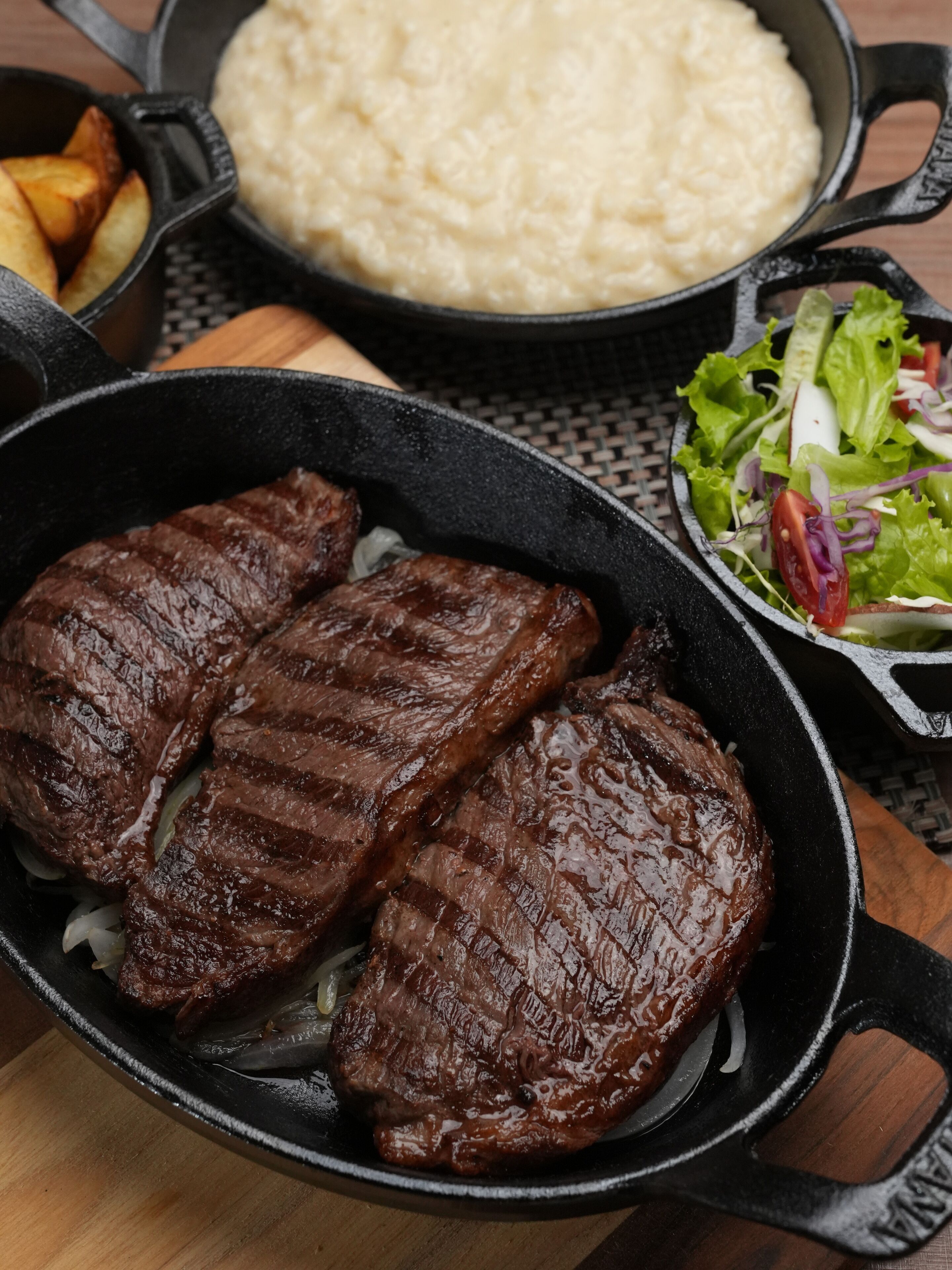 Une poêle contenant un steak grillé, un bol de riz et une salade, le tout posé sur une table en bois.