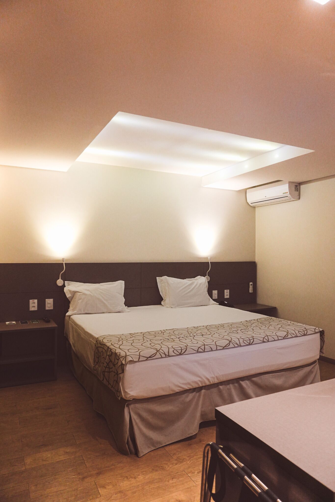 A hotel room with a bed, bedside tables, a desk, and an air conditioning unit.