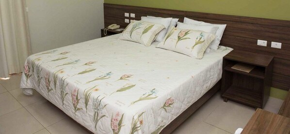 A neatly made bed with floral bedding, a wooden headboard, and a bedside table with a lamp. - Panorama Hotel (Juazeiro do Norte)