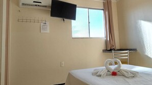 A hotel room with a bed, a flat-screen TV mounted on the wall, an air conditioning unit, a window with curtains, and a small desk with a chair. - Pousada Aconchego (Juazeiro do Norte)