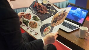 Une personne est assise à une table, devant un menu présentant différents plats et leurs prix, un ordinateur portable et une tasse blanche.