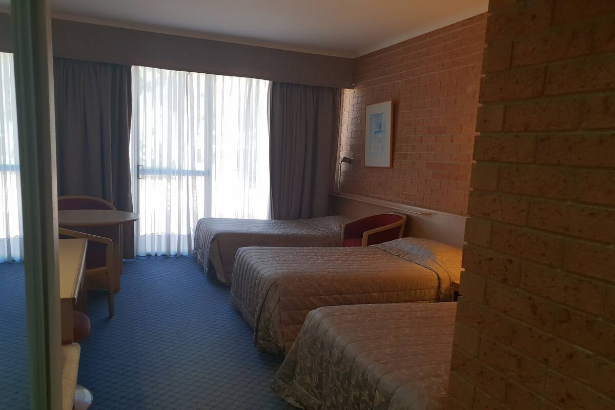 Ein Hotelzimmer mit zwei Betten, einem Schreibtisch und einem Stuhl. Es gibt ein Fenster mit Vorhängen, eine Ziegelwand und ein gerahmtes Bild an der Wand.