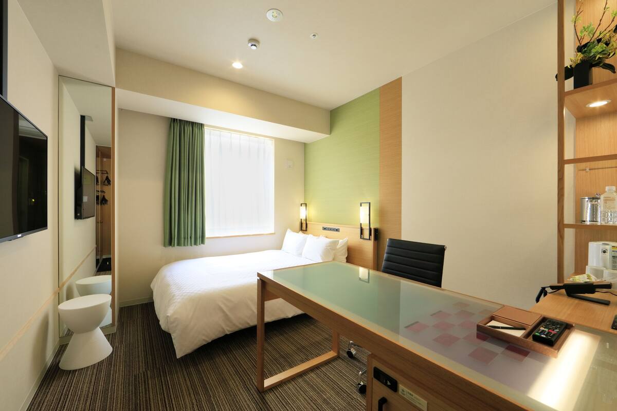 Habitación de hotel con cama, escritorio, silla, televisión, baño y estante con artículos de aseo.