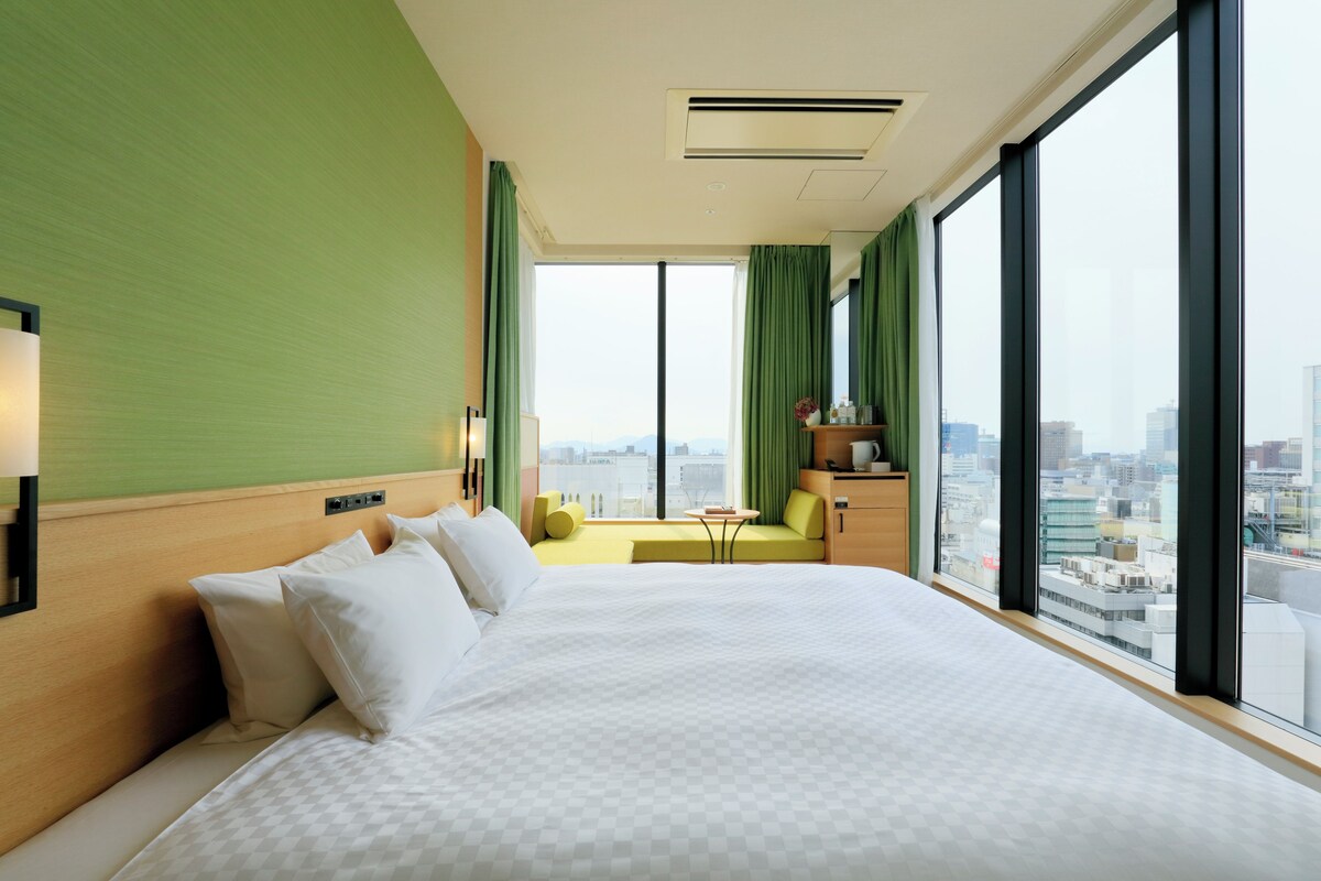 Une chambre d’hôtel avec un grand lit, une petite table, un canapé et une vue sur la ville grâce à de grandes fenêtres.
