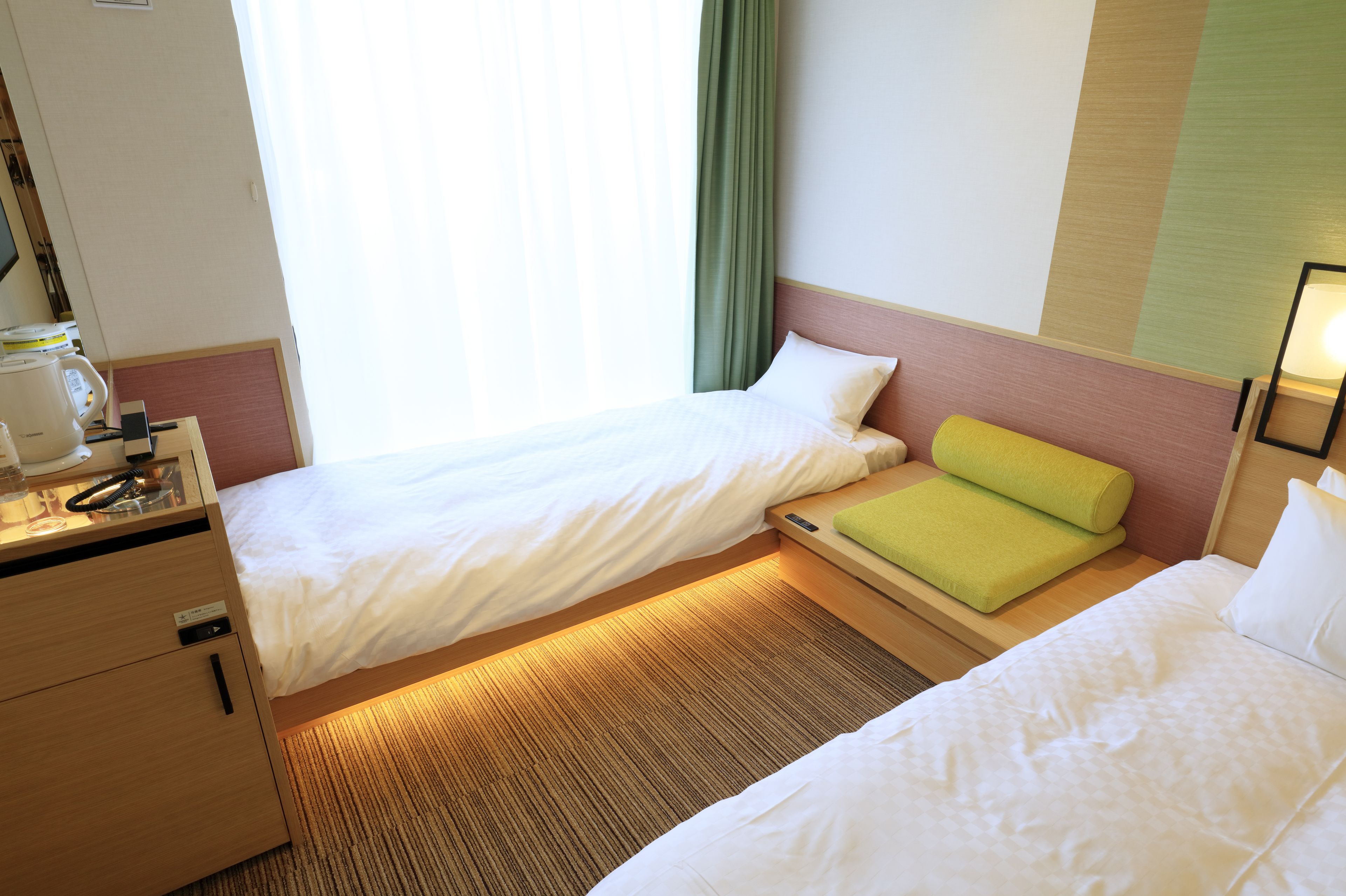 A hotel room with two single beds, a desk with a lamp, and a window with curtains.