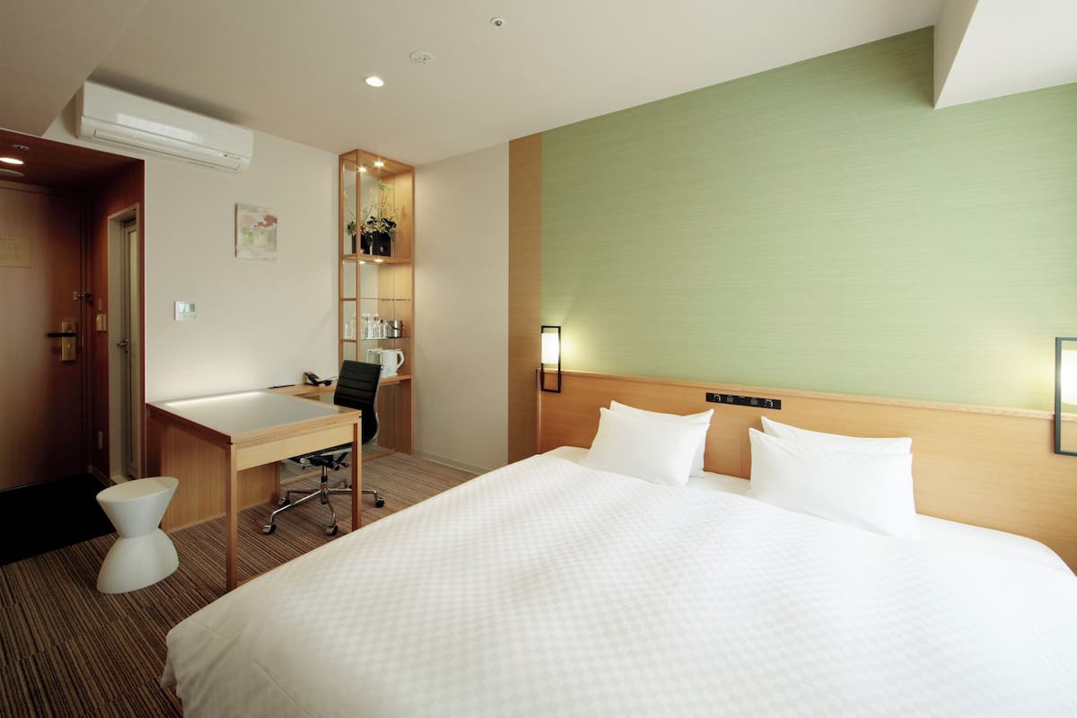 Habitación de hotel con una cama grande, un escritorio con una silla, un taburete pequeño, un estante en la pared con artículos de aseo y una lámpara de pared.