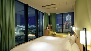 Habitación de hotel con una cama grande, una mesita de noche, una pequeña mesa y vistas a un paisaje urbano a través de amplias ventanas.