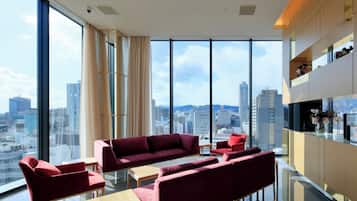 Una habitación de hotel moderna con ventanas amplias, un sofá rojo, una mesita pequeña y un mueble de madera con adornos.