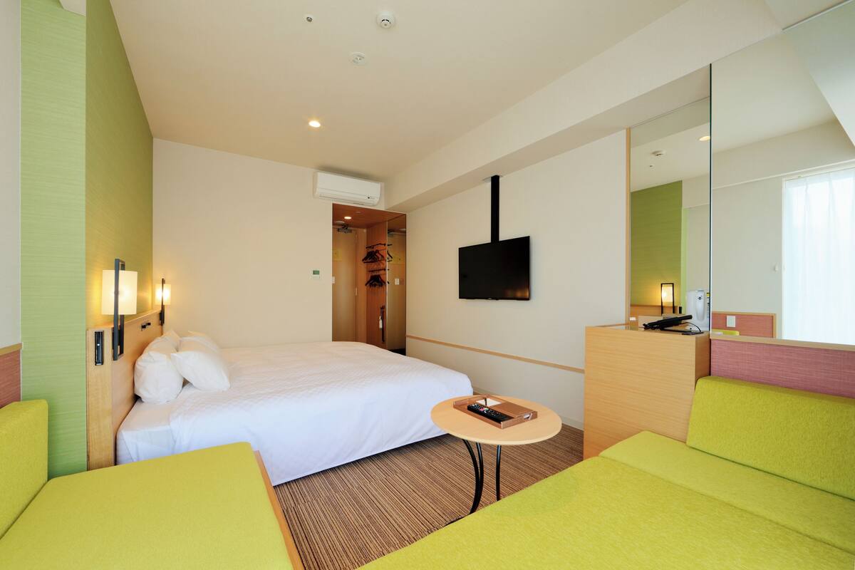 Habitación de hotel con cama, una pequeña mesa, un sofá, televisor montado en la pared y un espejo.