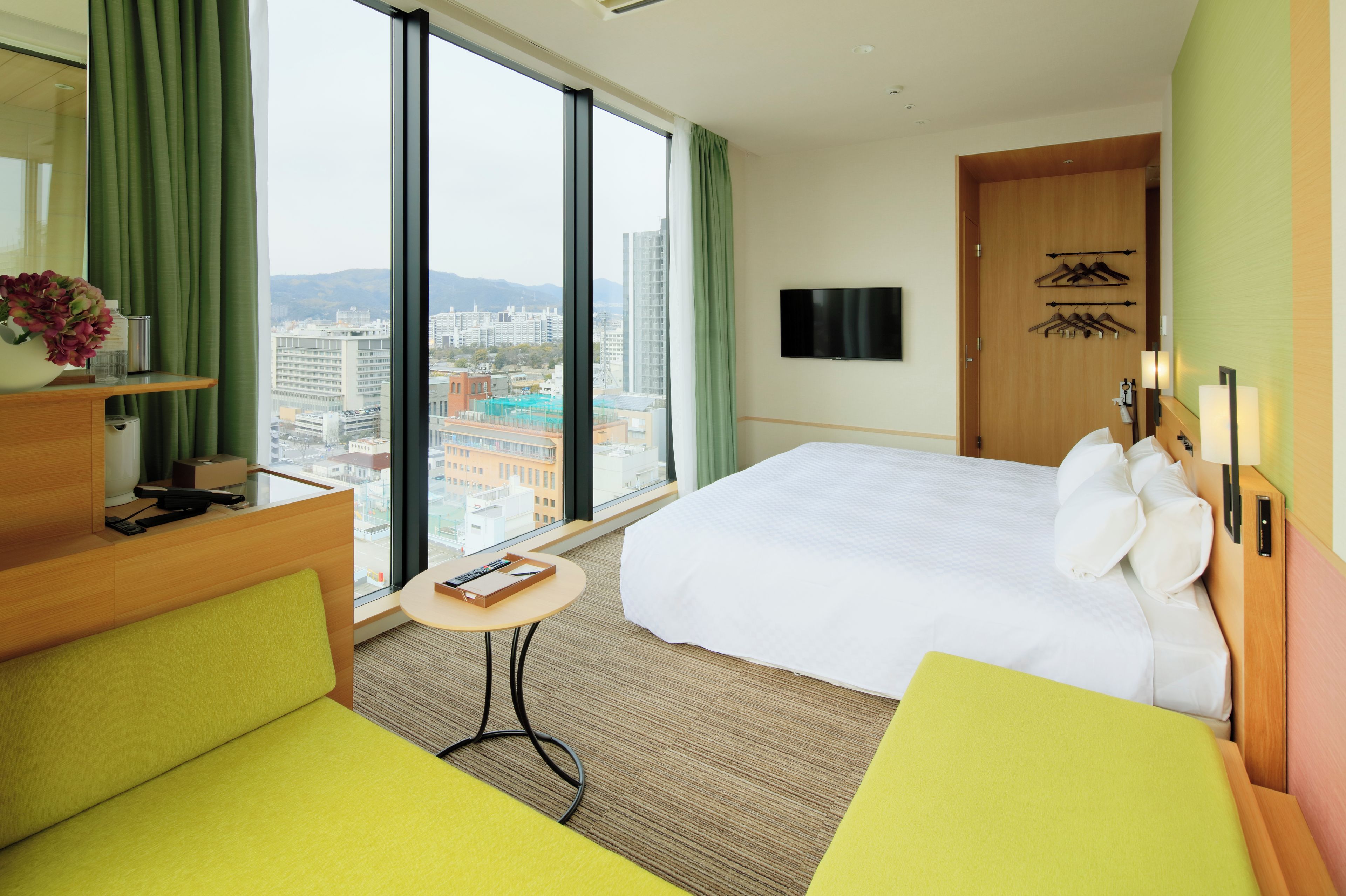 A hotel room with a large bed, a desk, a chair, a small table, a TV, and a view of the city.