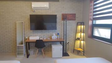 Habitación de hotel con pared de ladrillo, televisor de pantalla plana sobre un escritorio de madera, refrigerador compacto, silla de oficina negra, lámpara de pie y ventana con persianas.