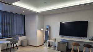 Habitación de hotel moderna con televisión de pantalla plana, escritorio, dos sillas, un refrigerador pequeño y una lámpara.