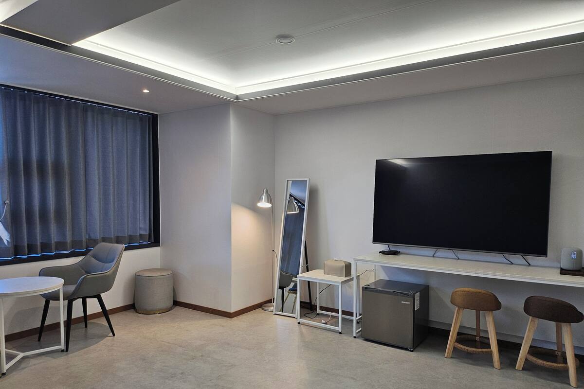 A modern hotel room with a flat-screen TV, a desk, two chairs, a small fridge, and a lamp.