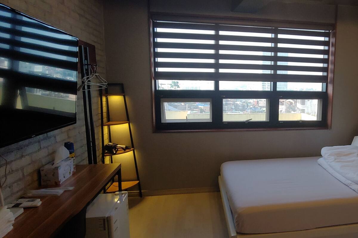 A bedroom with a bed, a TV, a desk, and a window with blinds.