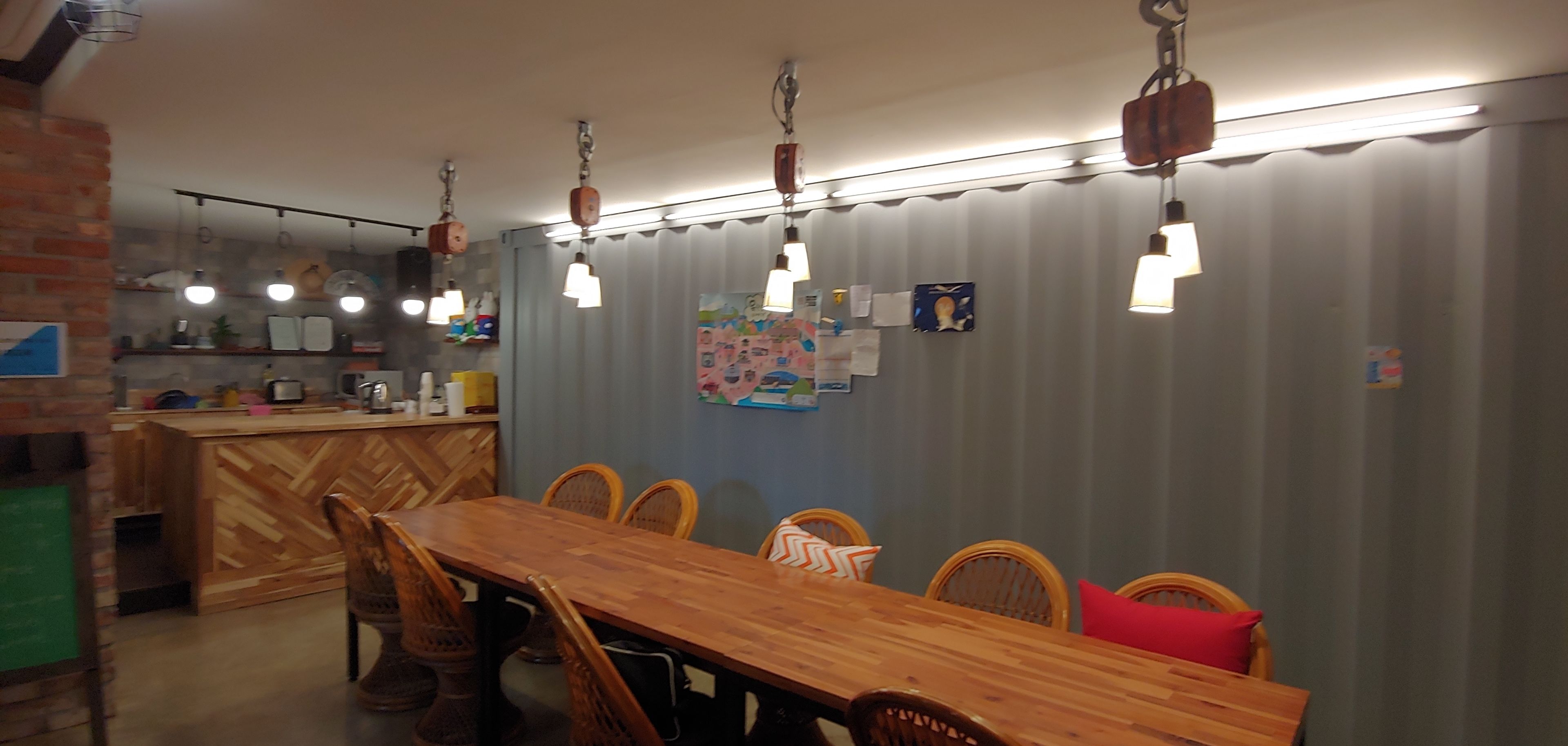 A dining area with a wooden table and chairs, a kitchen in the background, and hanging pendant lights.