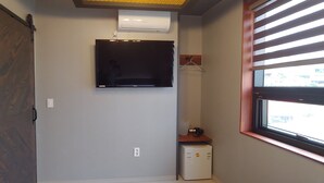 Une chambre dotée d’une porte coulissante, d’un climatiseur fixé au mur, d’un téléviseur à écran plat, d’un petit réfrigérateur et d’une fenêtre avec des stores.