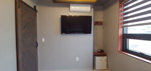 Una habitación con puerta corredera, aire acondicionado instalado, televisor de pantalla plana, refrigerador pequeño y ventana con persianas.