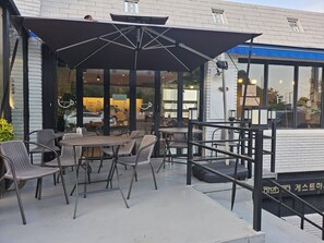 Un espace extérieur aménagé avec des tables et des chaises, une zone couverte avec des parasols, et un bâtiment doté de grandes fenêtres et d’une enseigne.