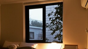 A bedroom with a bed, a bedside table with a lamp, and a window with a view of buildings and trees. - Hongdae Sopoong the Urban (Seoul)