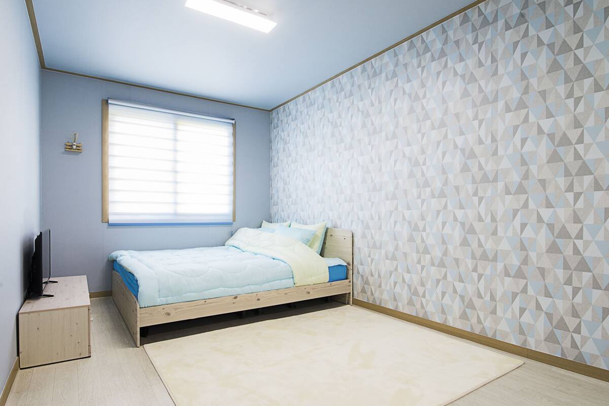 Une chambre à coucher avec un lit, une fenêtre, un mur orné d’un motif géométrique et une petite armoire en bois.