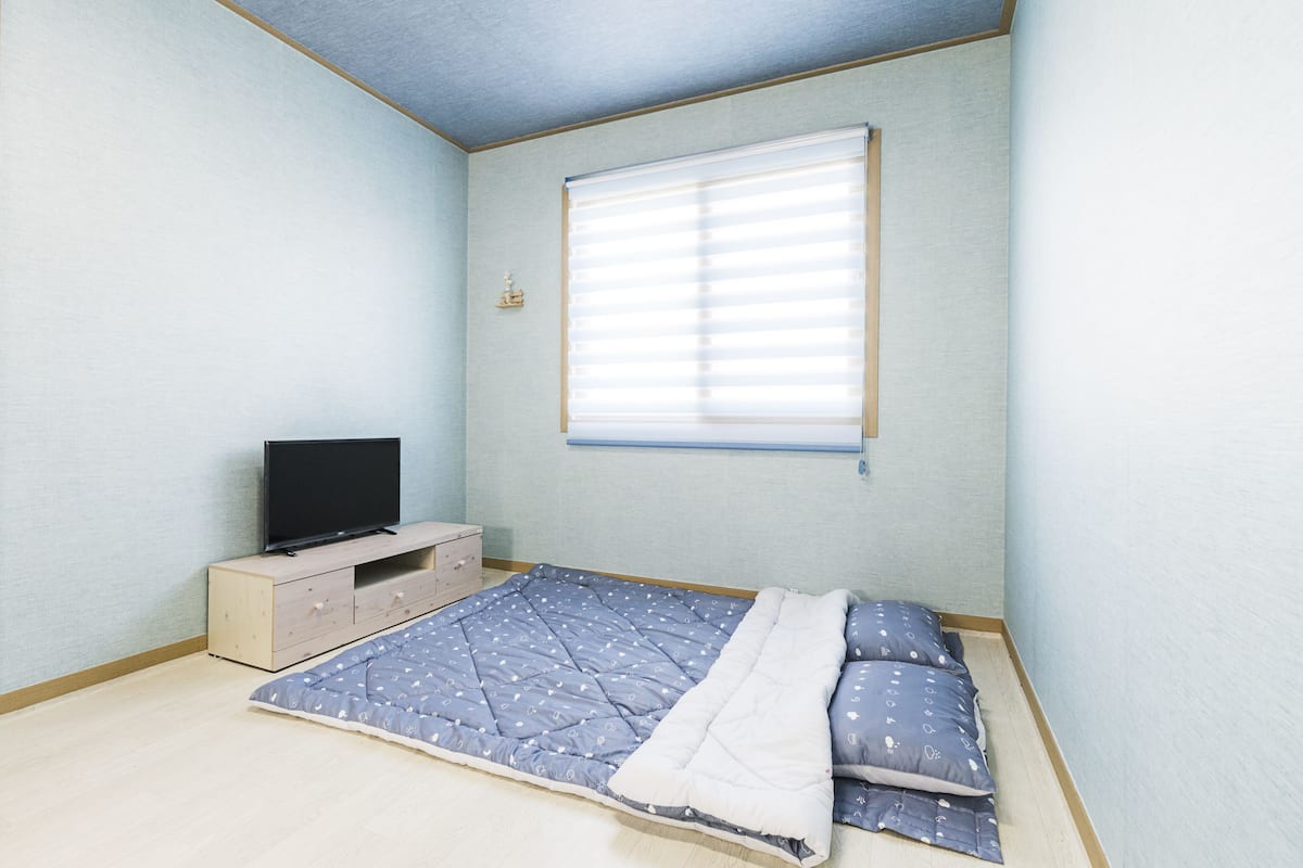 Une chambre à coucher avec un lit, un téléviseur posé sur une commode en bois et une fenêtre avec des stores.