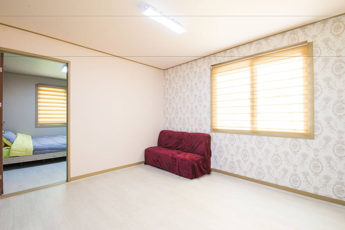 Une pièce avec un lit, un canapé rouge, une fenêtre avec des stores et un papier peint à motifs.