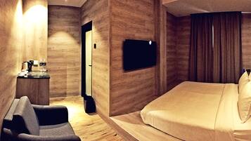 A modern hotel room with a bed, a seating area, a TV mounted on the wall, and a small table with a kettle and bottles.