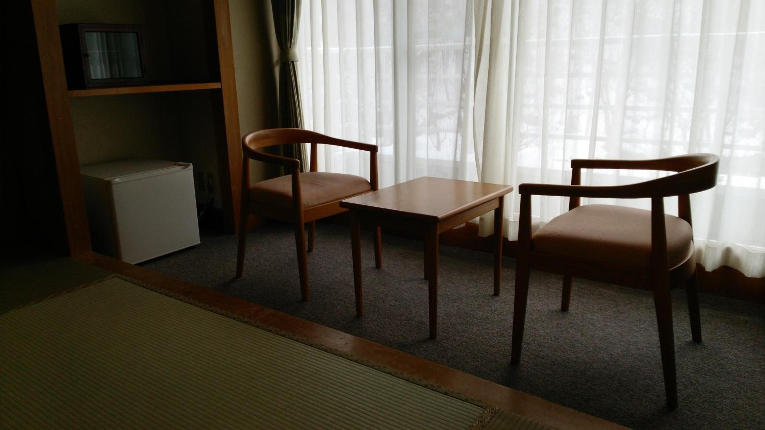Ein Zimmer mit einem kleinen, weißen Kühlschrank, zwei Holzstühlen, einem Holztisch und einem großen Fenster mit durchsichtigen Vorhängen.