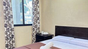 A neatly made bed with a pink bedspread and two rolled towels, a black nightstand with a cup and saucer, and a window with patterned curtains. - Pabua's Cottages (Mambajao)