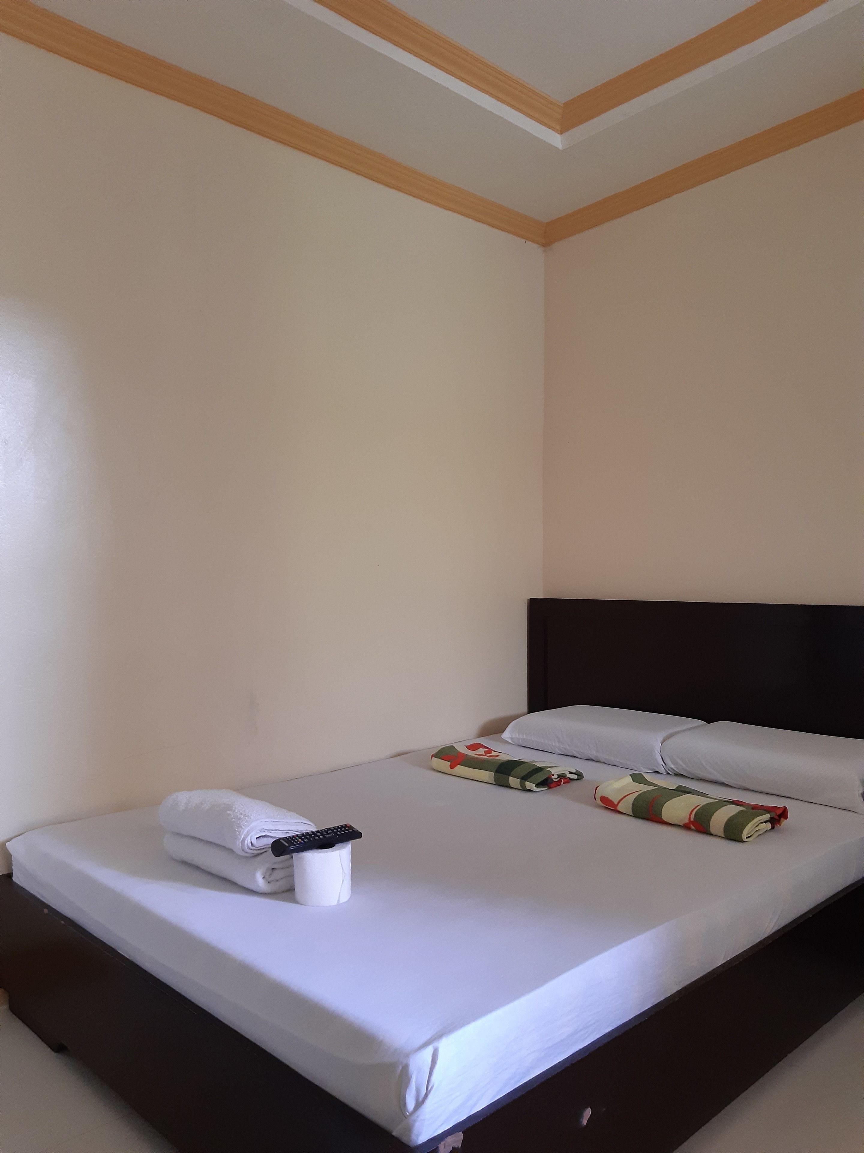Een net opgemaakt bed met witte lakens, een afstandsbediening en een opgerolde handdoek op een donker houten bedframe in een kamer met eenvoudige wanden en een houten plafondbalk.