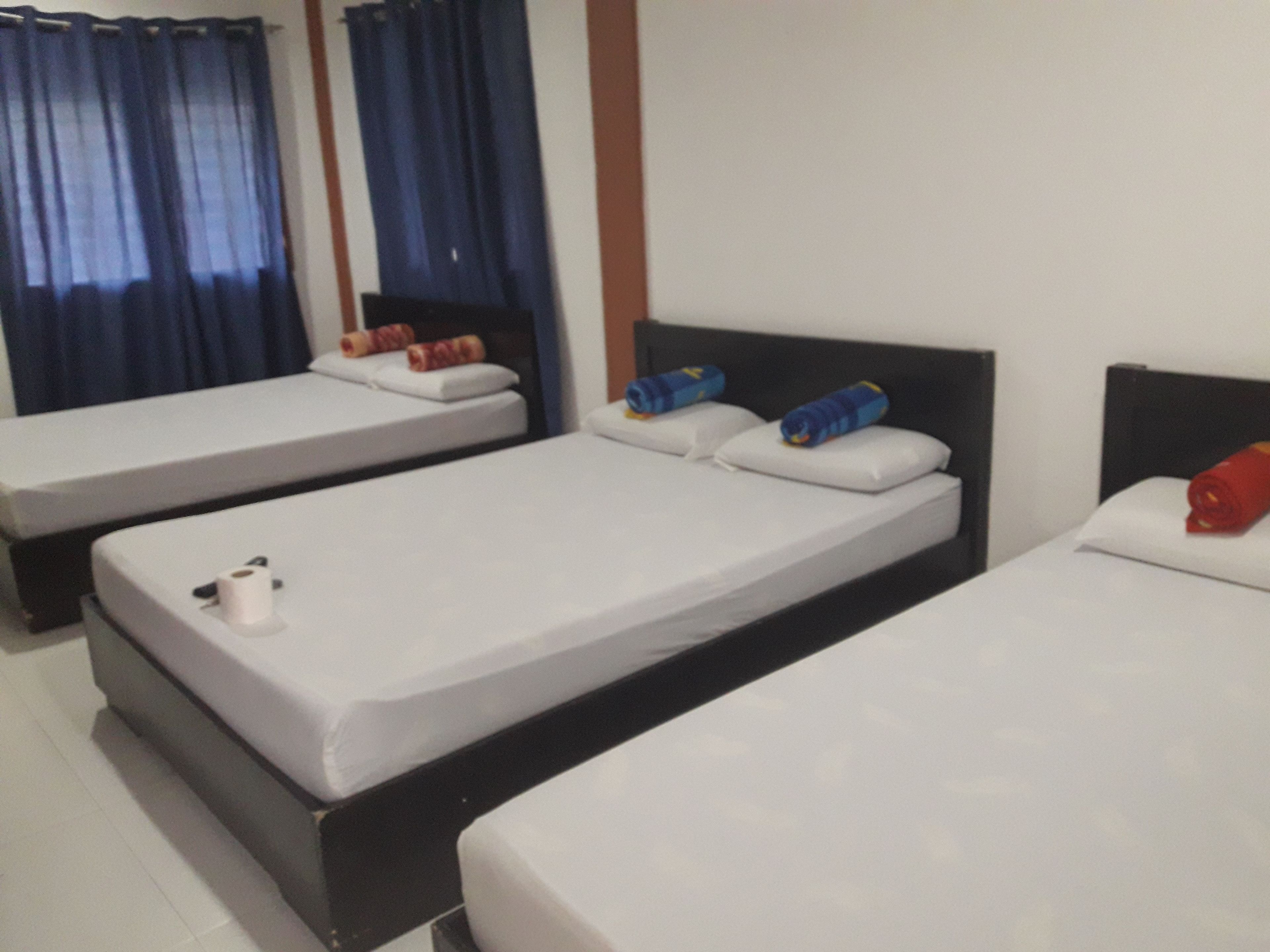 A hotel room with three single beds, each with white linens and pillows. There are towels and a bottle on the beds.
