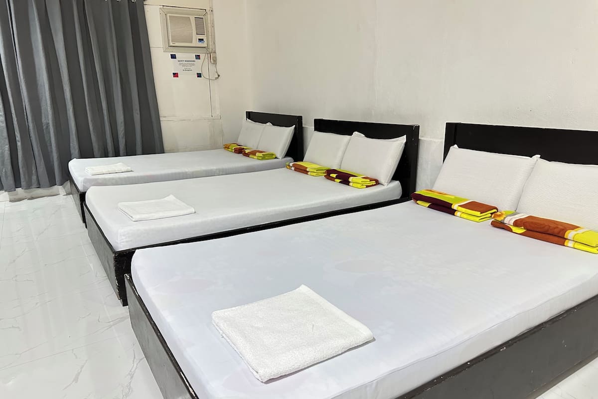 Quarto de hotel com três camas de solteiro, cada uma com colchão e travesseiro brancos. Há um aviso de segurança na parede e uma janela com cortinas cinzas.