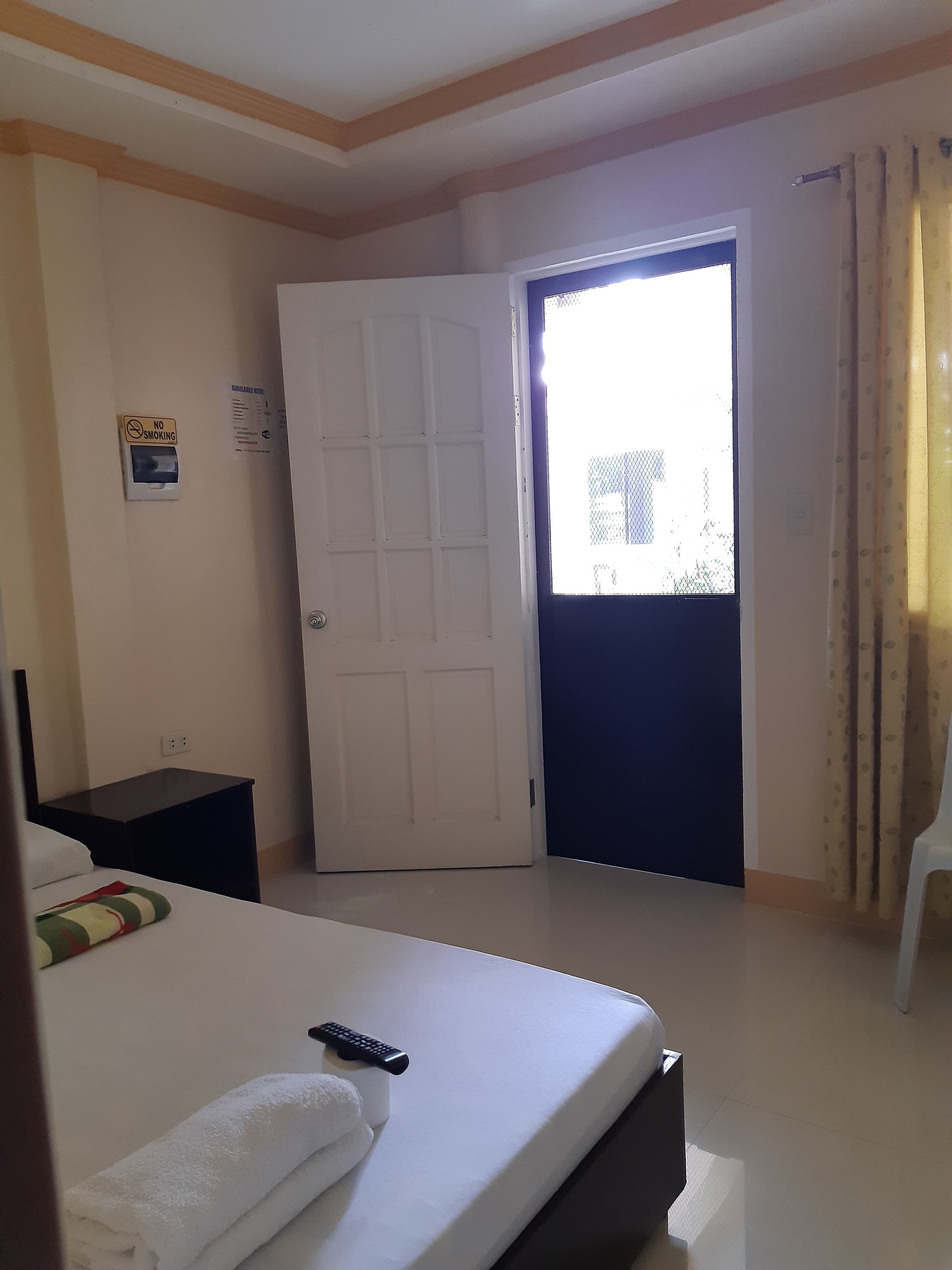 A hotel room with a bed, a bedside table, a remote control, a towel, a door, and a window with curtains.