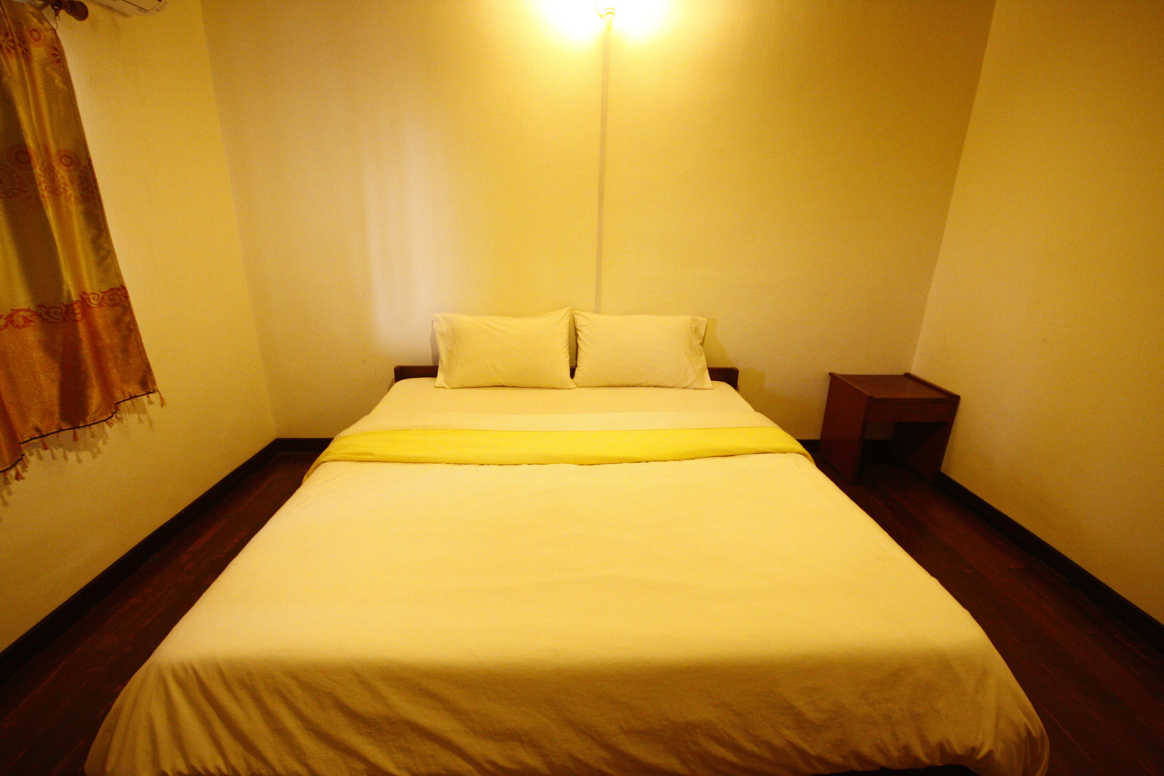 A neatly made bed with a yellow bedspread and pillows, a bedside table, and a wall-mounted light fixture.