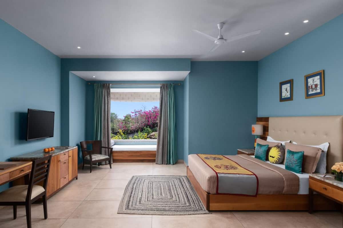 A bedroom with a bed, a desk, a chair, a TV, and a view of a garden.