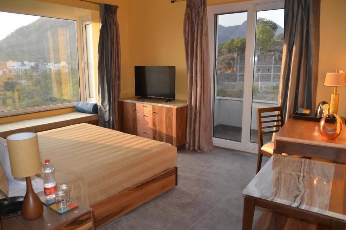 A hotel room with a bed, a TV, a desk with a lamp, a chair, and a view of the outside.