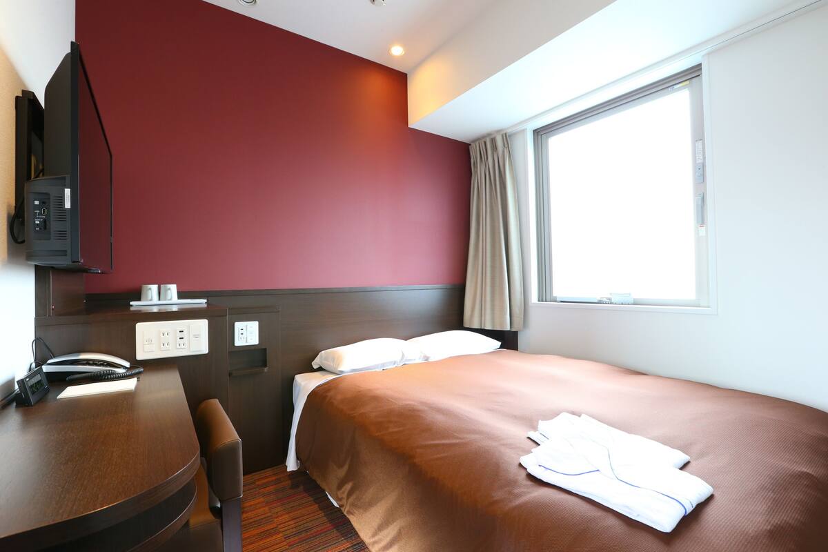 A hotel room with a bed, a desk with a telephone, a chair, a television, and a window with curtains.