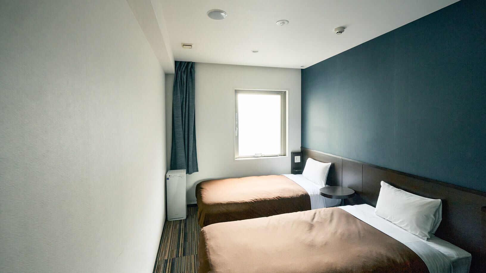 Hotellihuone, jossa on kaksi sänkyä, ikkuna, yöpöytä ja seinälle asennettu ilmastointilaite.