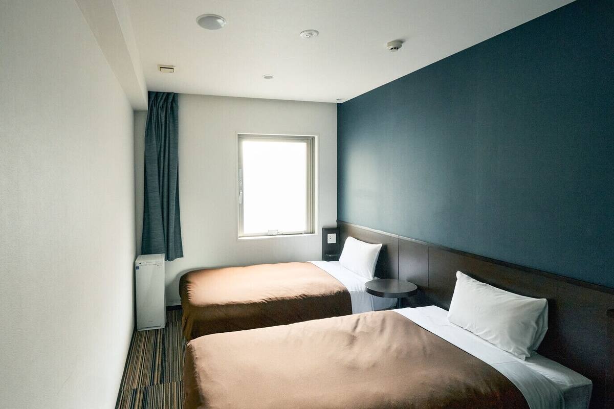A hotel room with two beds, a window, a bedside table, and a wall-mounted air conditioner.
