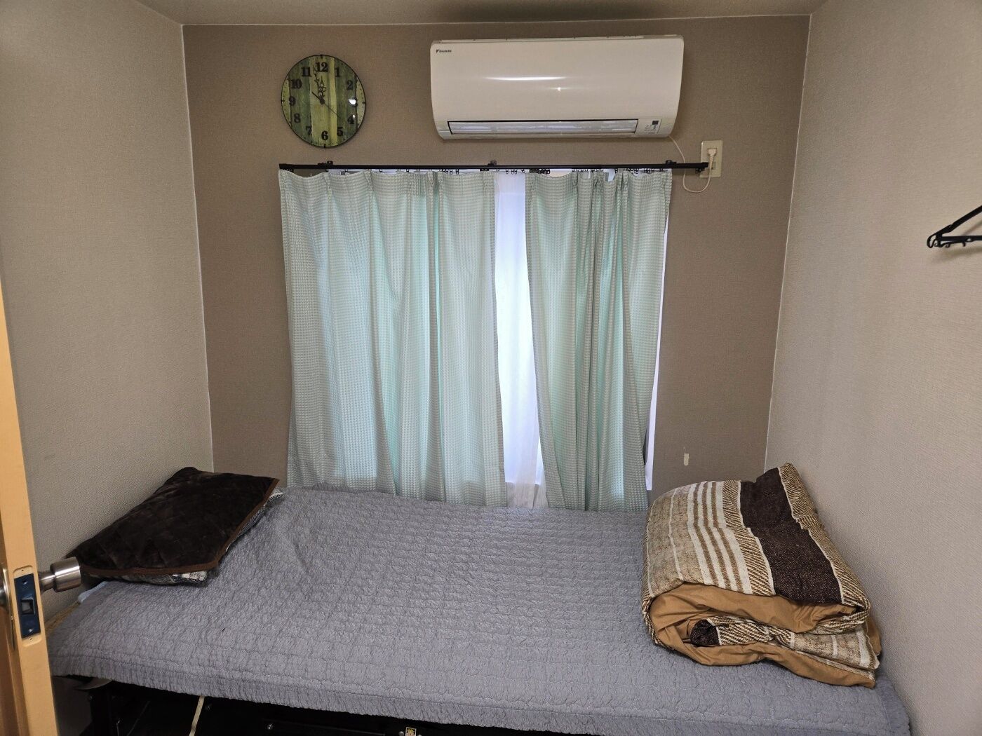 A small, single-bed room with a grey quilted bedspread, a brown pillow, a clock, and an air conditioning unit on the wall.