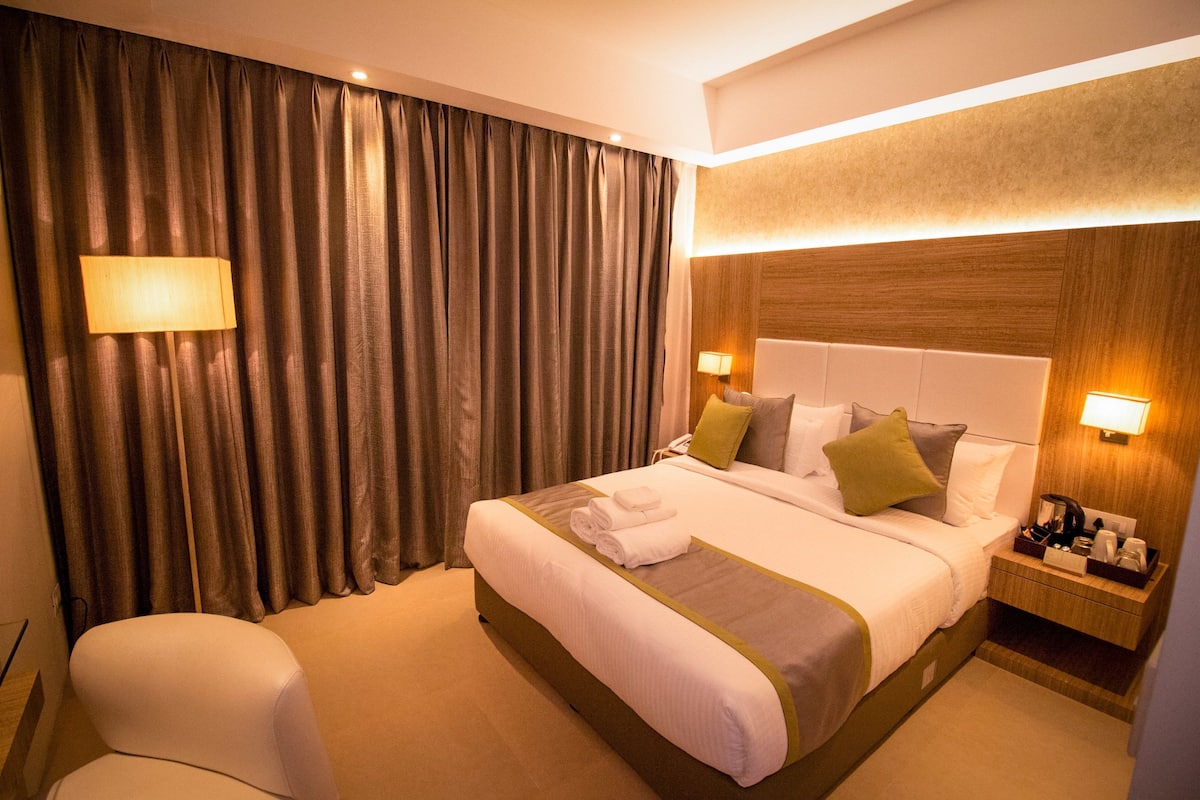 A hotel room with a large bed, a bedside table with a lamp, a chair, and a curtain.