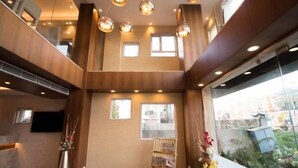 A hotel lobby with a high ceiling, wooden paneling, and a seating area with a glass wall providing a view of the outdoors. - The Maureen (Barasat)