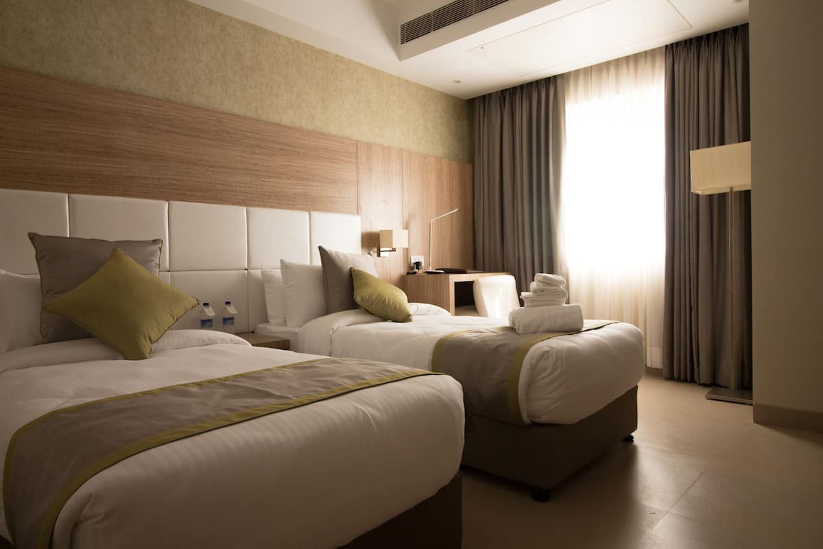 A hotel room with two beds, a nightstand, a lamp, and a window with curtains.