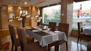 A dining area with a table set for four, surrounded by chairs, with a large window offering a view of buildings and a street outside. - The Maureen (Barasat)