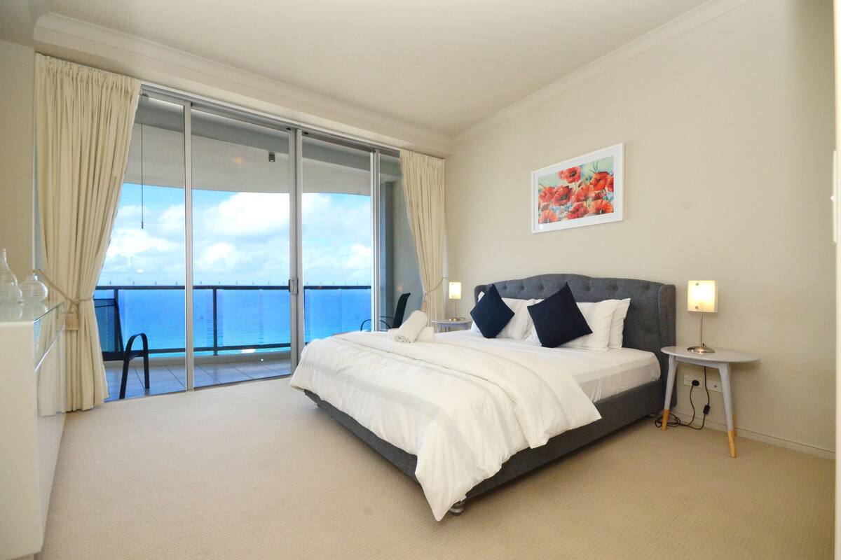 Une chambre d’hôtel avec un grand lit, une table de chevet, une chaise, un balcon avec vue sur la mer et un tableau au mur.