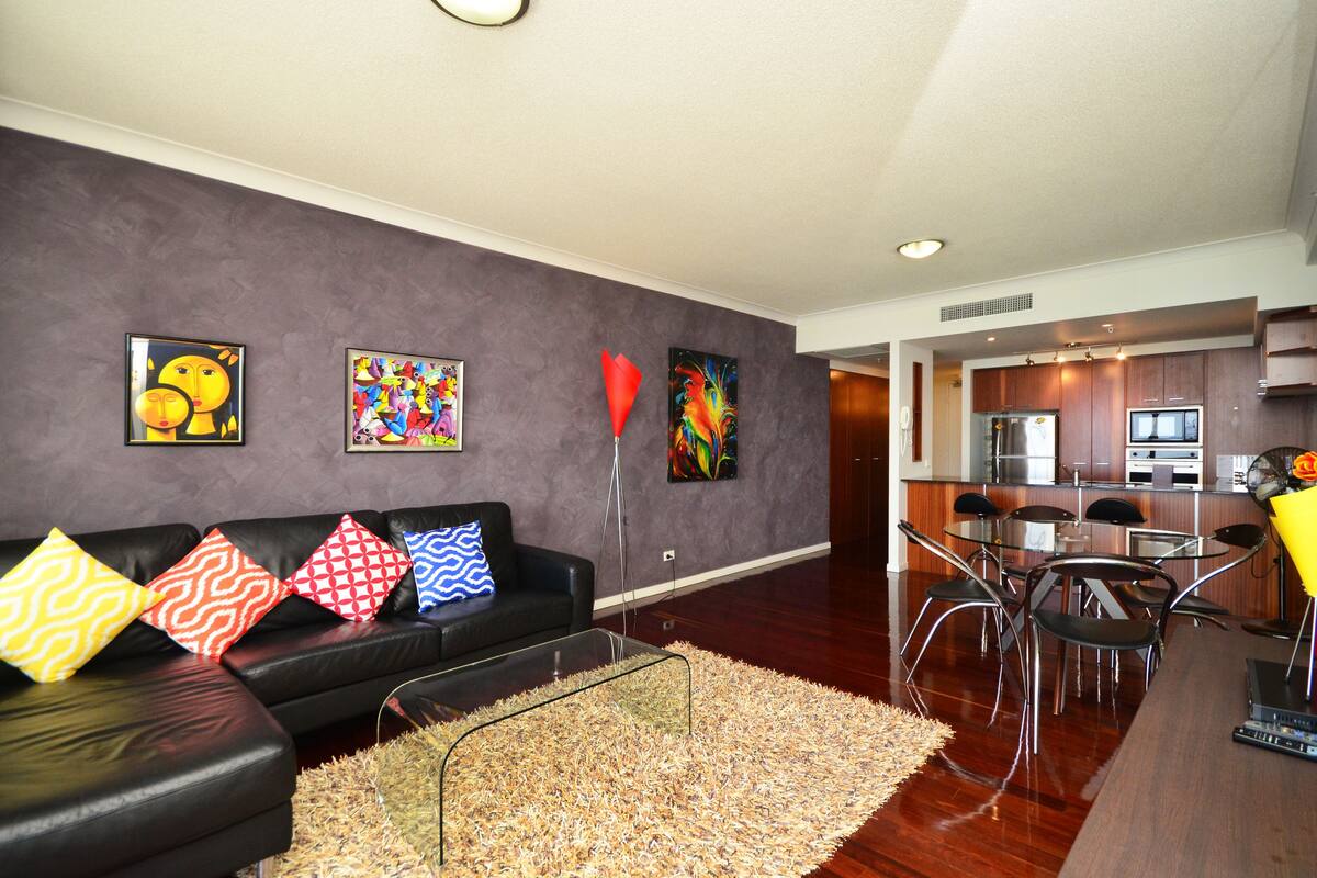Un salon moderne avec un canapé en cuir noir, des coussins colorés et des œuvres d’art abstraites accrochées au mur.