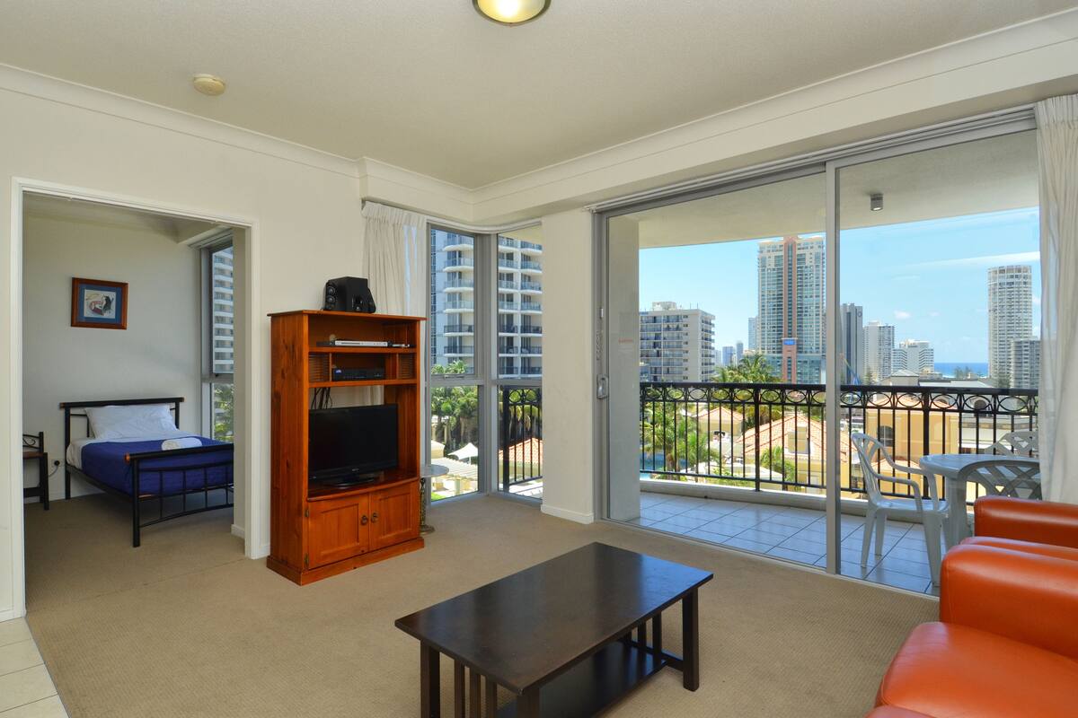 Un salon moderne comprenant un lit, un téléviseur, un meuble de rangement en bois et un balcon offrant une vue sur la ville.