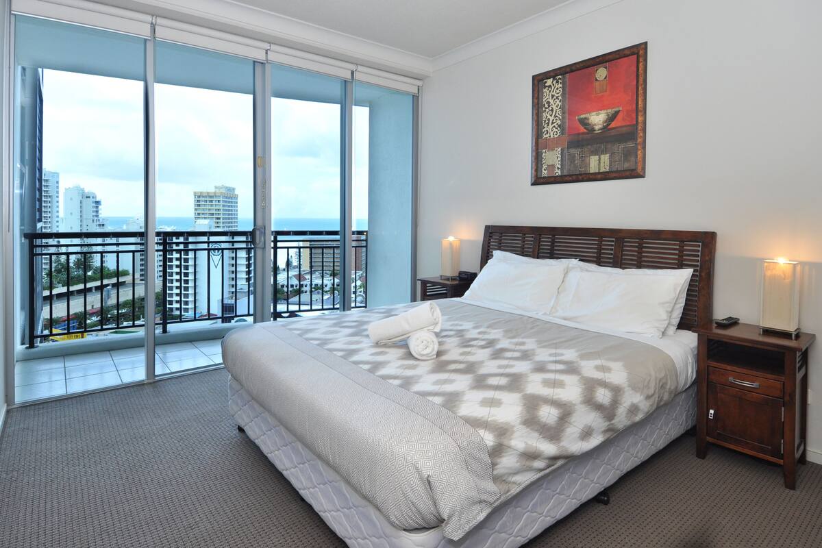 Une chambre d’hôtel avec un grand lit, une table de chevet, une lampe et une vue sur la ville grâce à de grandes fenêtres.