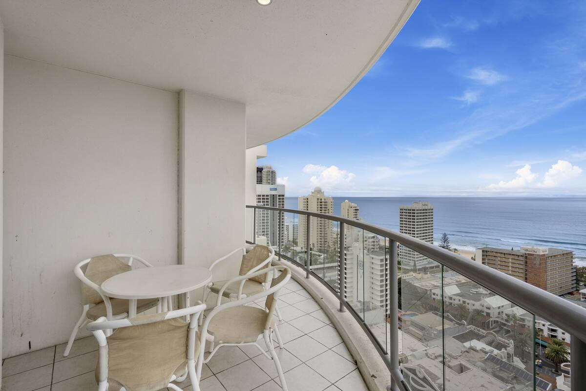 Un balcon avec des chaises blanches et une petite table, d’où l’on peut admirer le paysage urbain et l’océan.